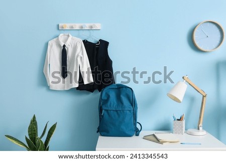 Desk with backpack, clock and stylish school uniform hanging on blue wall in room