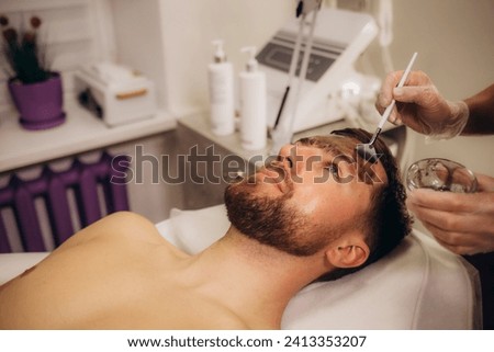 Side view of young serene man with closed eyes keeping head on rolled soft towel while female beautician applying clay mask on his face. High quality photo