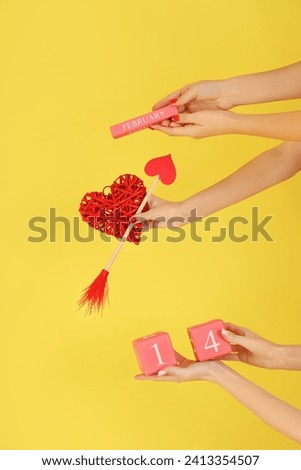 Female hands holding cube calendar with date 14 FEBRUARY, Cupid's arrow and heart on yellow background. Valentine's Day celebration Royalty-Free Stock Photo #2413354507