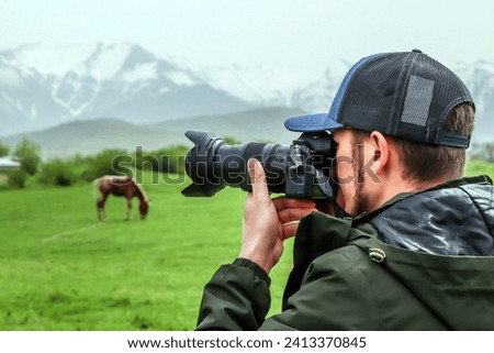 Photographer-naturalist.Photographer takes pictures with a camera standing on a green meadow with grazing horses against the backdrop of mountains