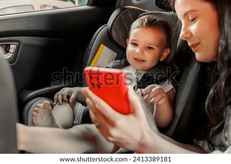 Mother and toddler son having fun while riding on back seat of the car, mother playing songs and cartoons to son using smart phone while baby is safely placed in car seat