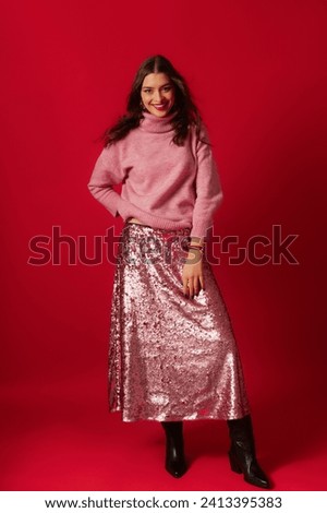 Fashionable beautiful happy smiling woman wearing trendy pink turtleneck sweater, midi sequin skirt, black leather cowboy boots, posing on red background. Full-length studio fashion portrait Royalty-Free Stock Photo #2413395383