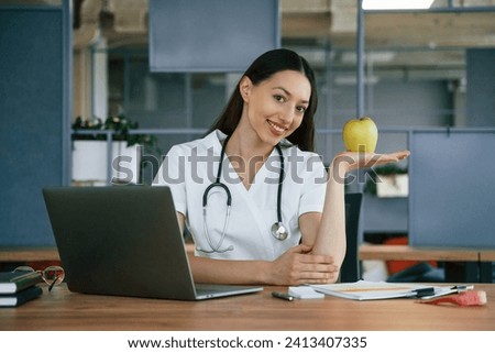 Green apple and laptop on table. Female doctor nutritionist in white coat is indoors.
