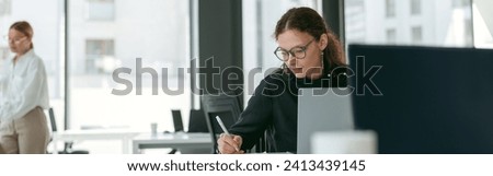 Stylish businesswoman making notes while working on laptop in modern office on colleagues background