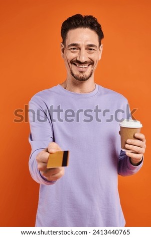 happy man in purple sweater holding coffee to go and offering credit card on orange background