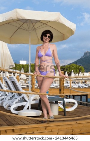 Woman 40-44 years old in a lilac bikini under a sun umbrella on the beach. Royalty-Free Stock Photo #2413446465