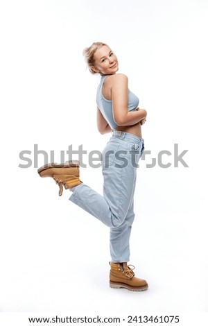 A smiling young girl stands tall. Cute blonde girl in a blue top, jeans and yellow boots. Side view. White background. Vertical. Full height. 