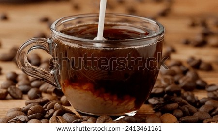 A stream of milk pours into a full cup of coffee and dissolves into it. Close up of a glass coffee or tea cup on the kitchen table. A morning ritual, a breakfast concept.