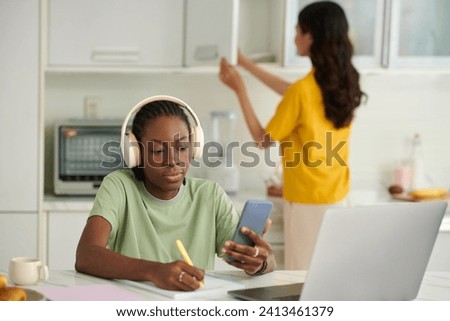 College student watching lection on smartphone and taking notes, when roommate cooking lunch Royalty-Free Stock Photo #2413461379