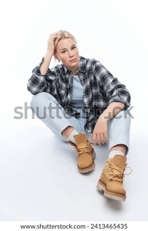 A pensive young girl is sitting on the floor. Cute blonde girl in jeans, plaid shirt and yellow shoes. White background. Vertical.