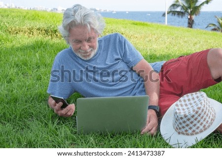 Senior bearded man lying down in the meadow close to the sea using laptop and mobile phone. Smart work concept in open air Royalty-Free Stock Photo #2413473387