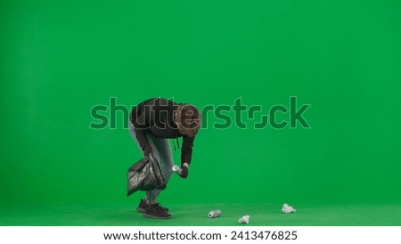 A male volunteer wearing black gloves and carrying a trash bag picks up trash on the green screen. The man puts paper and tin cans into the garbage bag. Voluntary free work assistance help concept.