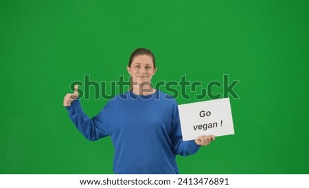 Woman holds a placard that says go vegan and shows a thumbs up. Portrait of female activist in a studio on a green screen close up.
