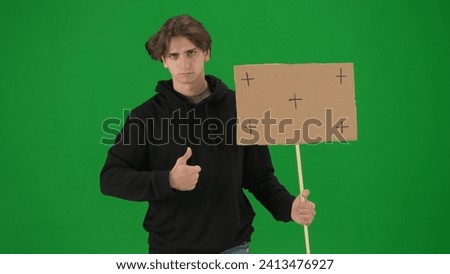 A male activist holds a blank poster and shows a thumbs up. A young man in casual clothes holds a placard on a green screen in close up. Chromakey, advertisement, promo.