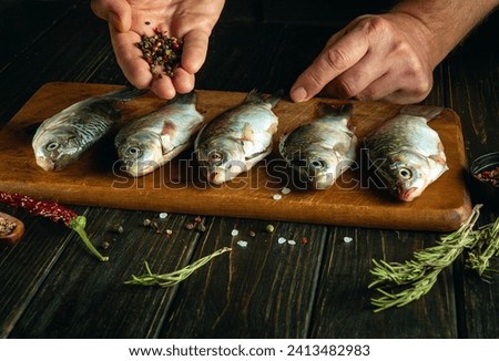 The cook prepares crucian carp fish with aromatic spices and dry pepper on a cutting board. The concept of cooking small carp on the kitchen table. Royalty-Free Stock Photo #2413482983