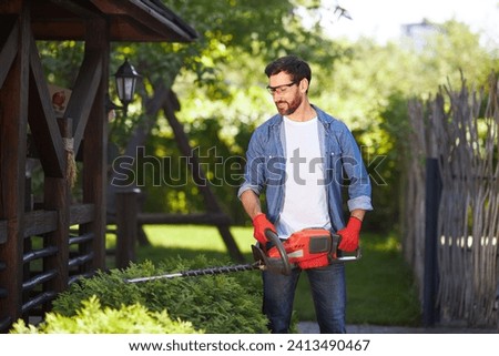 Professional landscaper in safety gloves cutting evergreen hedge with trimmer in park. Front view of enjoyed caucasian man pruning conifer bush with electric lopper and smiling. Concept of work. Royalty-Free Stock Photo #2413490467