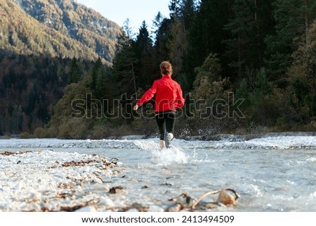 Rear View of a Female Curly hair Trekker Running Carefree On and Over an Alpine River Stream in Mountain Vacations in a Beautiful Afternoon Light