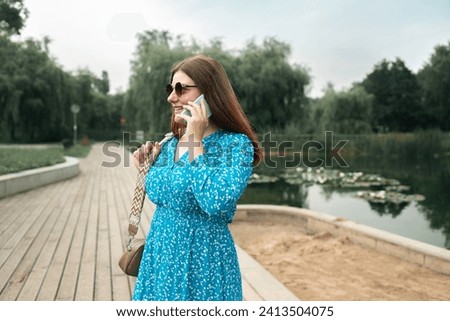 Young pretty woman model in stylish dresses talk by mobile cell phone look aside standing on summer in spring park outdoors. Urban lifestyle concept. High quality photo Royalty-Free Stock Photo #2413504075
