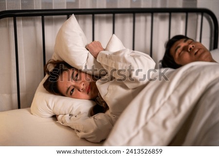 Asian woman feel frustrated from husband snoring while sleep at night. Attractive insomnia angry wife lying down on bed, cannot sleep due to noise and put cozy blanket on her ears in bedroom at home. Royalty-Free Stock Photo #2413526859