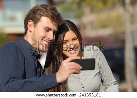 Happy couple watching media in a smart phone outdoors with an urban background