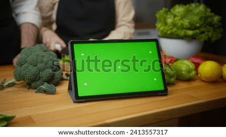Tablet computer device with mock up green screen chroma key display on the kitchen counter, married spouses chopping greens for salad, meal