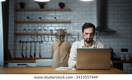 Young man is working on his laptop in the kitchen at home while his wife working in kitchen