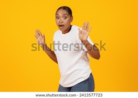 Confused frightened African American lady got caught posing with raised hands looking at camera with scared expression, standing over yellow studio background. It's not me concept Royalty-Free Stock Photo #2413577723
