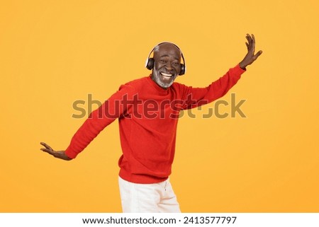 Joyful happy elderly African American man in a red sweater and headphones, dancing with his arms outstretched, displaying happiness and rhythm on a bright yellow background Royalty-Free Stock Photo #2413577797