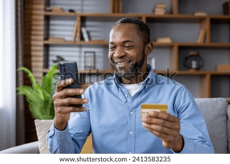 A smiling African American man is at home, sitting on the sofa, holding a credit card and using a mobile phone. Close-up photo.