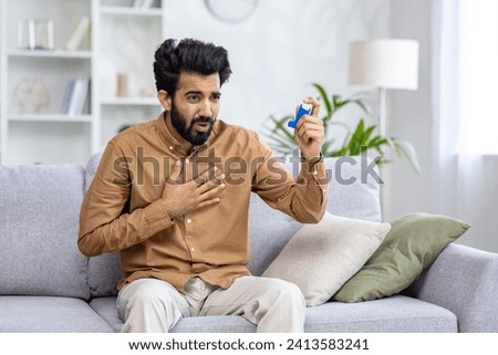 A young Indian man suffers from asthma and allergies. Sitting at home on the couch, holding his chest with his hand and using an aerosol medicine. Royalty-Free Stock Photo #2413583241