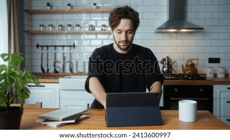 A young curly hair man sitting in kitchen, using app with tablet computer, surfing on internet