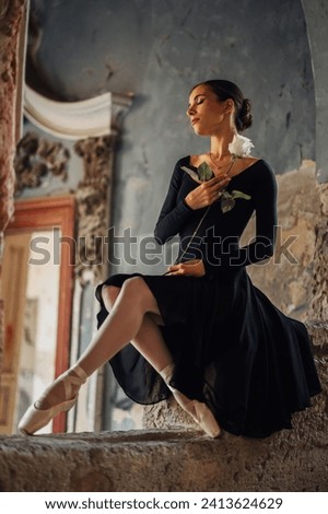 An elegant and gracious ballerina in black skirt is sitting on a rustic wall in abandoned castle with a white rose in her hands. Portrait of a lonely ballerina finding peace at abandoned castle. Royalty-Free Stock Photo #2413624629