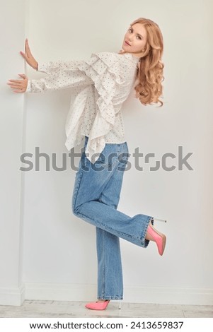Feminine beauty and fashion. Full-length portrait of a pretty blonde girl with delicate pink makeup posing in an elegant white blouse and blye jeans on a white studio background. Hairstyles, wave.