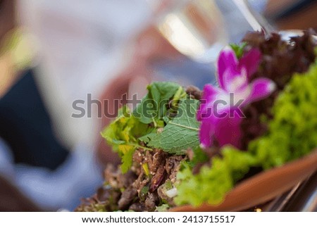 The photograph is a close-up of a gourmet salad presented in a terracotta bowl, garnished with a vibrant, purple edible flower. The salad consists of various greens, including frilly lettuce and herbs Royalty-Free Stock Photo #2413715517