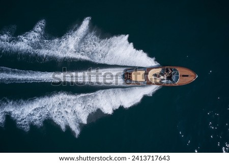 A large modern high-speed wooden luxury boat moves on blue water, top view. Expensive wooden boat, man and woman in motion on the water making a white trail looking like air. Royalty-Free Stock Photo #2413717643