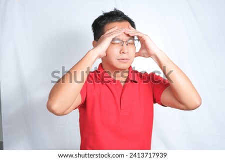 A young Asian man wearing red shirt and glasses is feeling dizzy or stressed. Isolated white background.