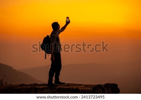 people and sun concept, man hold smartphone on the cliff of the mountain on sunset background, tourist travel in nature for trip, sun background and silhouette sky, man wearing backpack and take photo Royalty-Free Stock Photo #2413723495