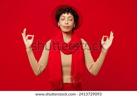 Young woman of African American ethnicity wears beige sweater hat scarf hold spreading hands in yoga om aum gesture relax meditate try to calm down isolated on plain red background. Lifestyle concept