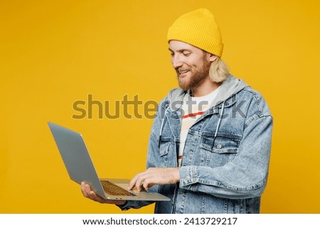 Side view young blond IT man wears denim shirt hoody beanie hat casual clothes hold use work on laptop pc computer typing message isolated on plain yellow background studio portrait. Lifestyle concept