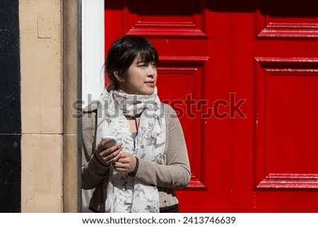 Portrait of a happy Chinese woman standing by a red door holding a smartphone. Lifestyle image.