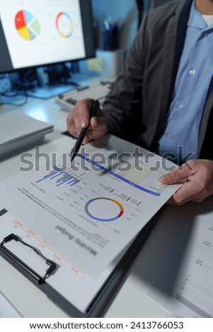 Young Asian businessman holds a pen and points to a chart graph on a document to analyze market data, finance, balance sheet, net profit, taxes to plan an online sales strategy startup business idea.