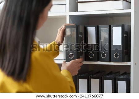 Attractive Asian woman in a yellow suit stands and pulls files from a shelf looking for financial documents in a meeting. Presentation within the company Management concept.
