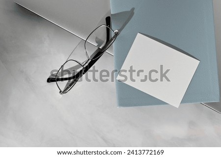 Empty paper business card mockup, blue notebook, eyeglasses, laptop on marble gray table background. Minimal aesthetic business workspace.