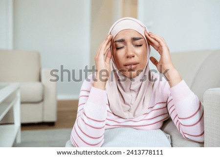 Portrait of a young Arabic Muslim woman sitting on the couch at home with a headache and pain. Woman with hijab suffering from chronic daily headaches. Sad woman holding her head because sinus pain