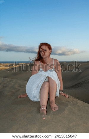 A young pretty red-haired girl in a white dress sits in the sand dunes in the late afternoon and enjoys the last rays of sunshine