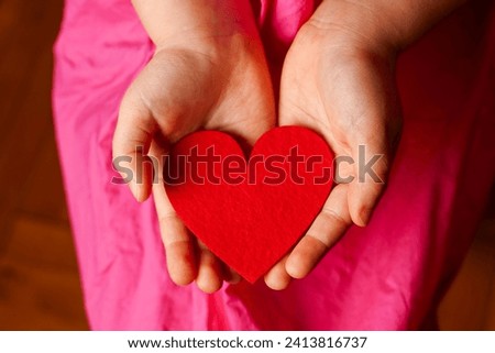 Child's hands holding bright red heart on pink background Royalty-Free Stock Photo #2413816737