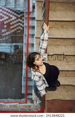 The girl is standing inside an industrial or residential building. A landing, an unkempt building. Women's clothing style is free