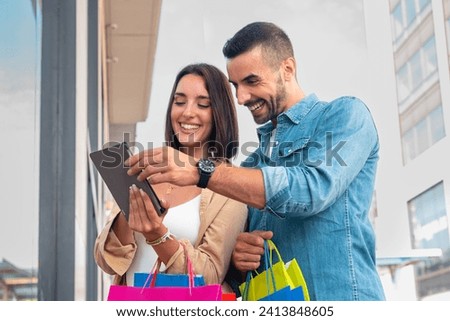 Smiling young Caucasian couple holding shopping bags, browsing tablet apps