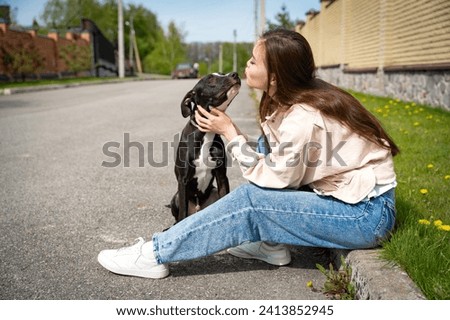 Heartwarming image of a young girl sitting on the curb, sharing a tender moment as she kisses her affectionate pitbull breed dog. The vibrant spring weather enhances the sweetness of the scene.