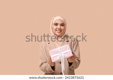 Happy young Muslim woman in hijab with gift box on beige background